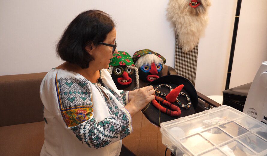 Romanian artisan Ana Cazac sewing a handmade ritual mask with a red nose and bean teeth, surrounded by other colorful folk masks in her workshop.