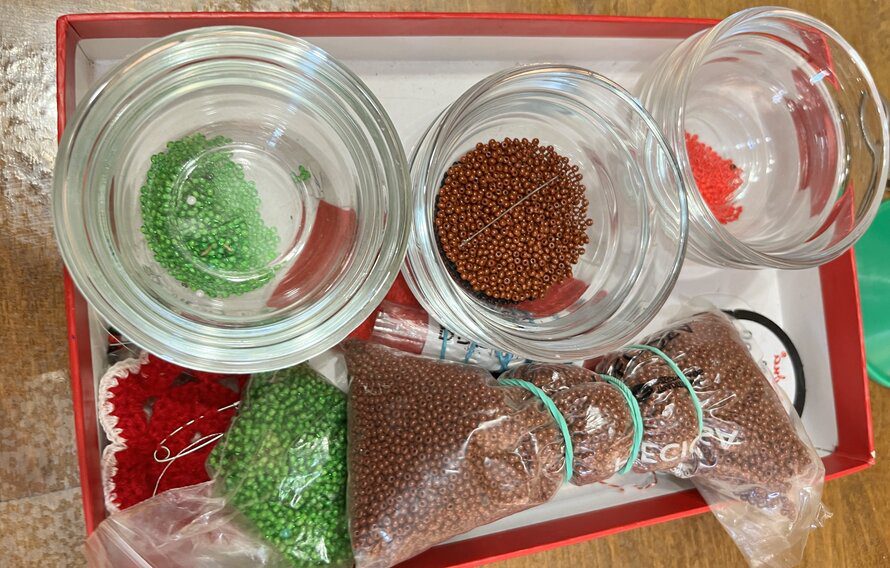 A collection of small glass bowls and plastic bags filled with colorful seed beads in green, brown, and red shades, arranged neatly in a box for beadwork art projects.