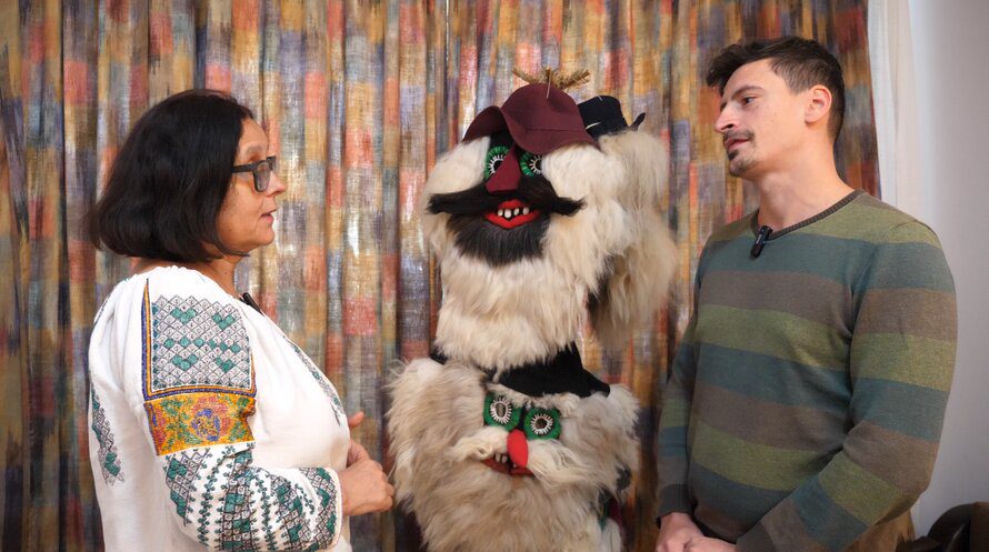 Author Catalin Geangos talking with Romanian artisan Ana Cazac, with two large traditional ritual masks made of wool, fur, and beans displayed between them.