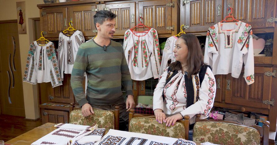 Catalin Geangos and artisan Viorica in a room with traditional Romanian beaded shirts displayed on hangers behind them, discussing beadwork craft.