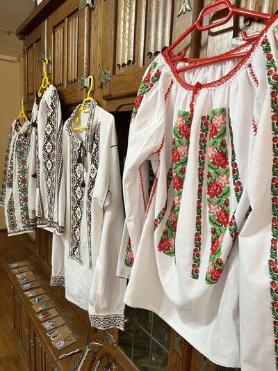 White Romanian blouses with colorful beadwork patterns, including floral and geometric designs, hanging on wooden cabinets.