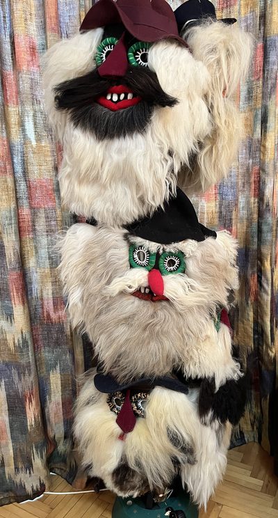 Handmade Romanian folk masks stacked vertically, featuring wool beards, fur details, bean teeth, and bright red noses, displayed against patterned curtains.