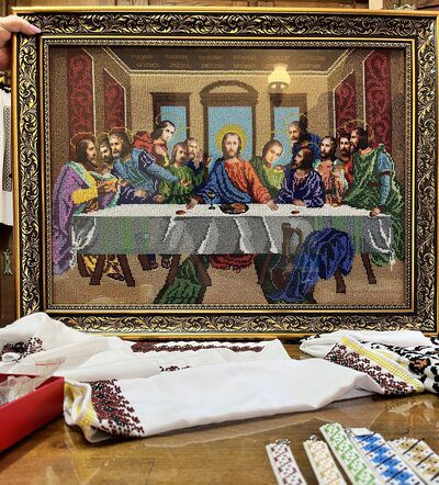 A framed bead embroidery depicting The Last Supper, created with colorful seed beads, shown alongside traditional embroidered fabrics and beadwork materials.