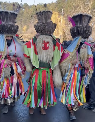 Three Romanian men wearing traditional ritual masks with fur, red noses, tall feathered hats, and multicolored costumes during a folk celebration parade.