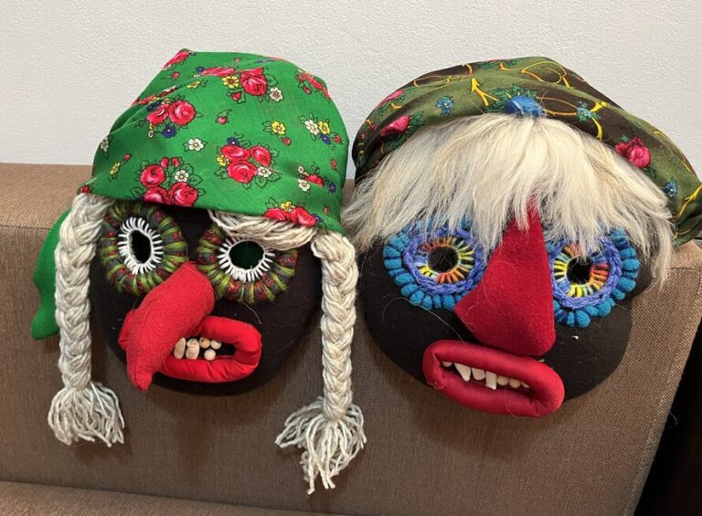 Two traditional Romanian ritual masks resting on a couch, featuring red noses, bean teeth, wool braids, and floral headscarves, crafted for village performances.