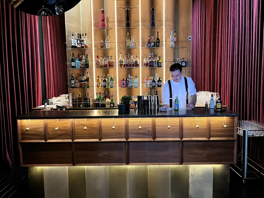 Bartender behind a wooden cocktail bar with illuminated bottle shelves and burgundy curtains