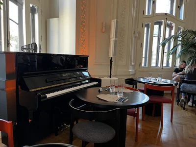 Black upright piano beside tables and tall windows in the historic salon