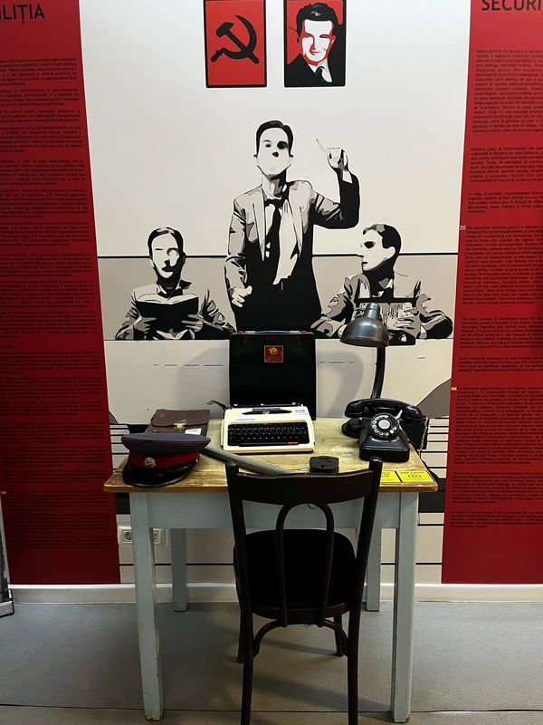Militia and Securitate panels behind a desk with a typewriter and rotary telephone.