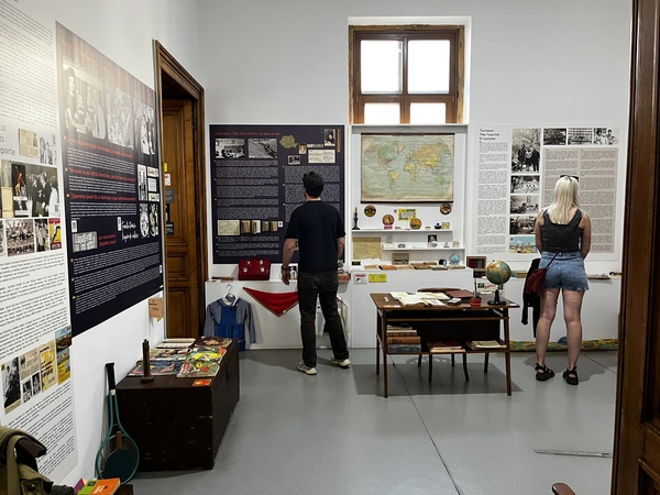 Museum visitors beside a school desk, wall map and everyday-life displays.